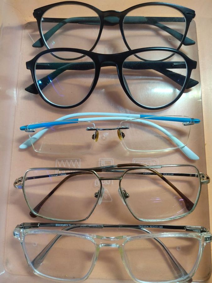 Five pairs of used glasses stacked in a clear tray: two black round frames, a rimless pair, a metal frame and a clear acetate frame